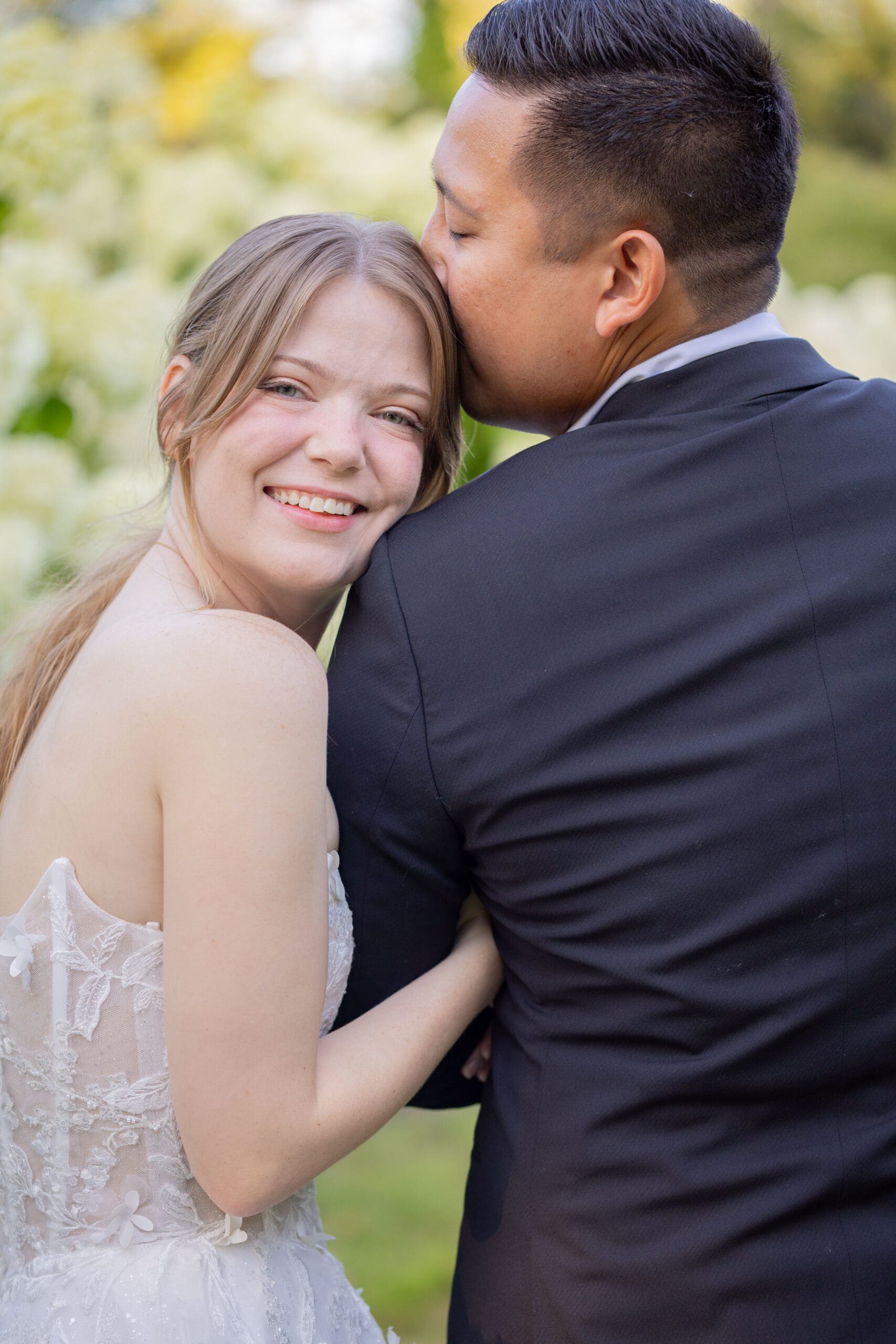 groom-kissing-brides-forehead-while-bride-looks-at-camera-smiling
